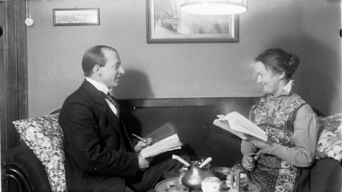 En mann og en kvinne sitter i en sofa og ser p? hverandre og smiler, mens de holder hver sin bok.